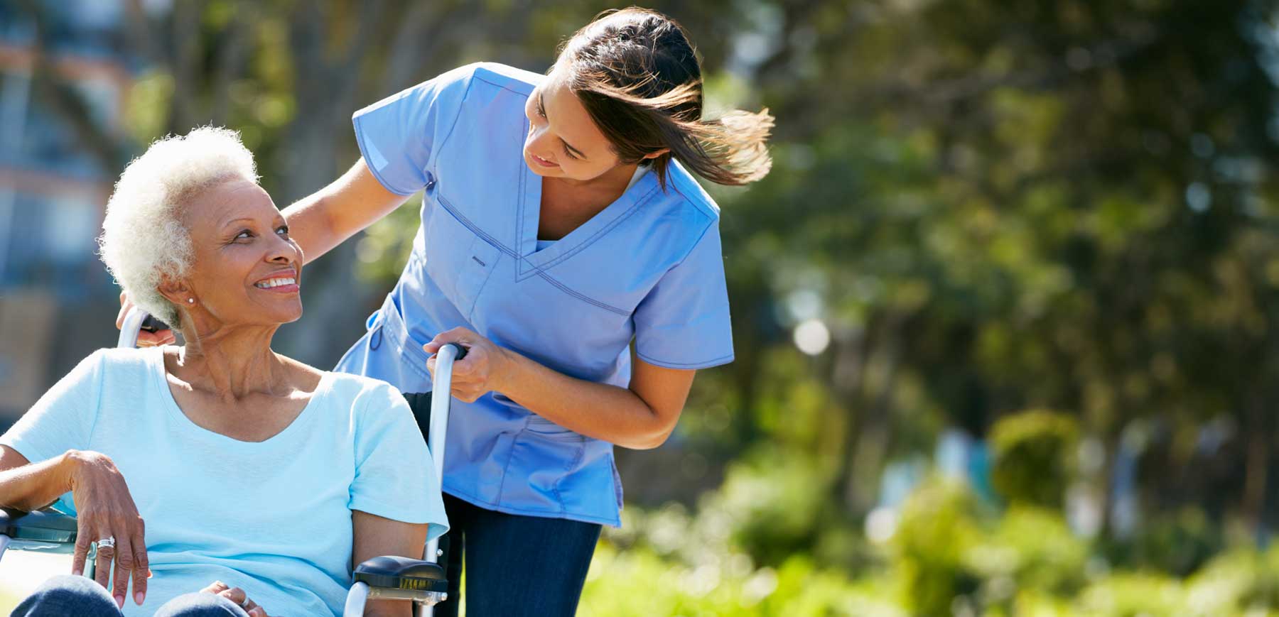 A smiling caregiver gently pushes an elderly woman in a wheelchair outdoors on a sunny day, sharing a warm moment together in a peaceful, green park-like setting, reflecting compassionate, personalized care.
