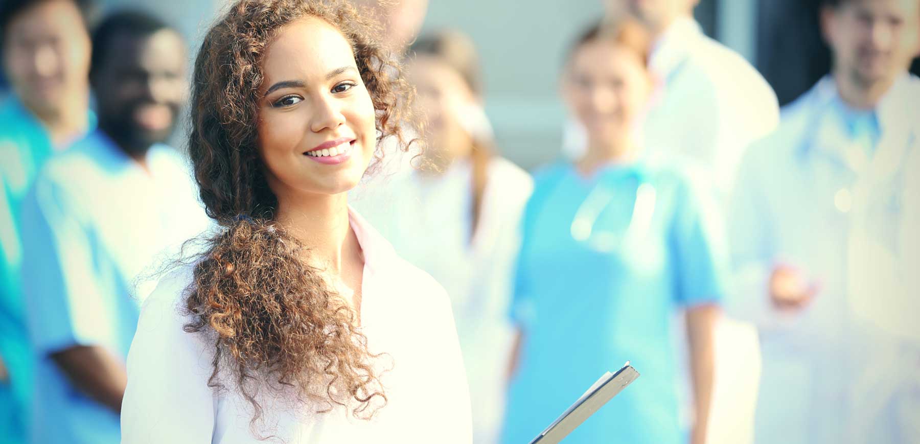 Smiling healthcare worker holding a clipboard stands confidently in the foreground, with a diverse team of nurses, aides, and medical professionals blurred in the background, reflecting teamwork and career opportunities in home care and nursing.