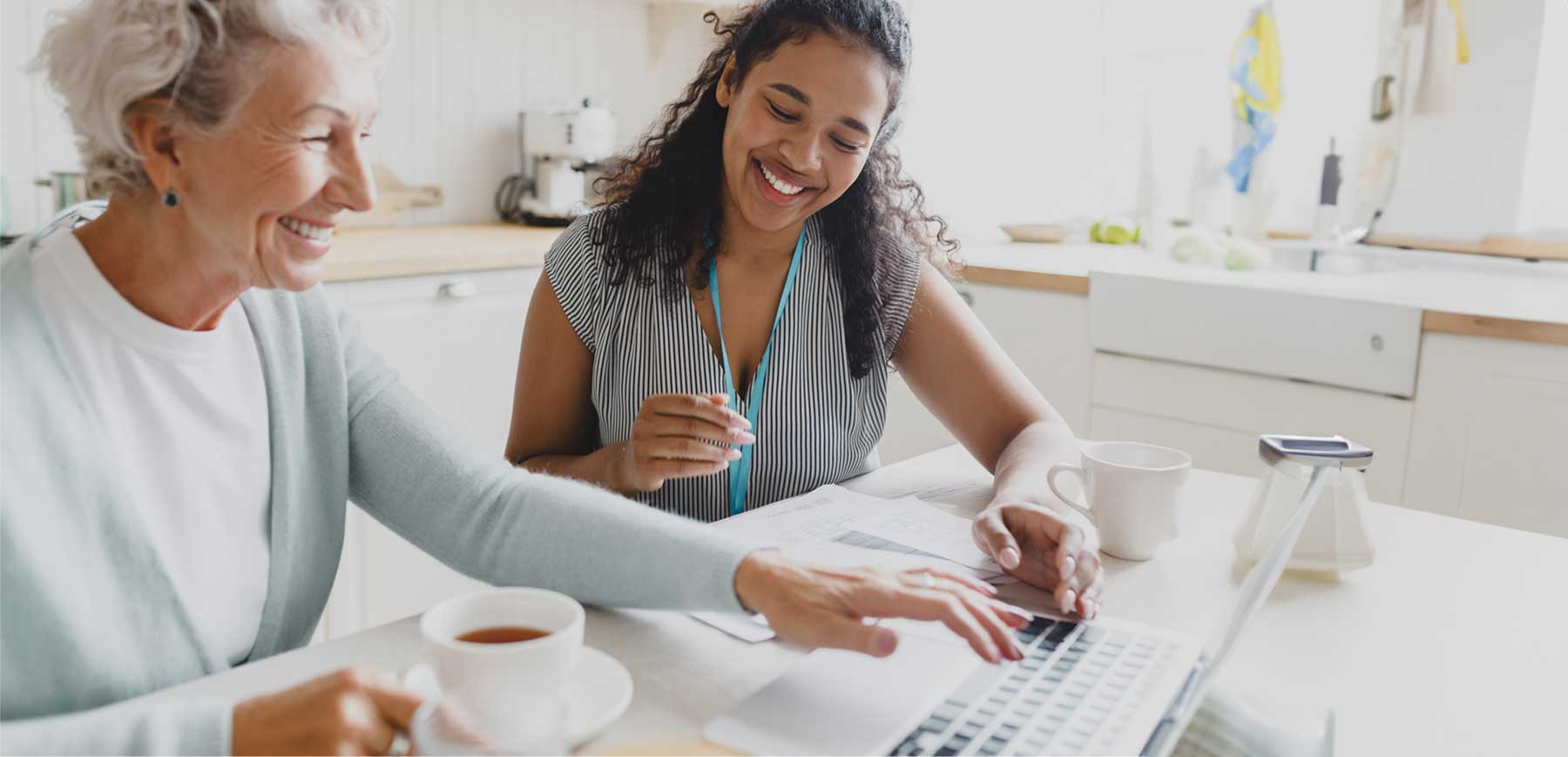 Smiling caregiver sits with an older woman at a kitchen table, reviewing paperwork and using a laptop together over coffee, reflecting personalized assistance, supportive care, and friendly client communication.
