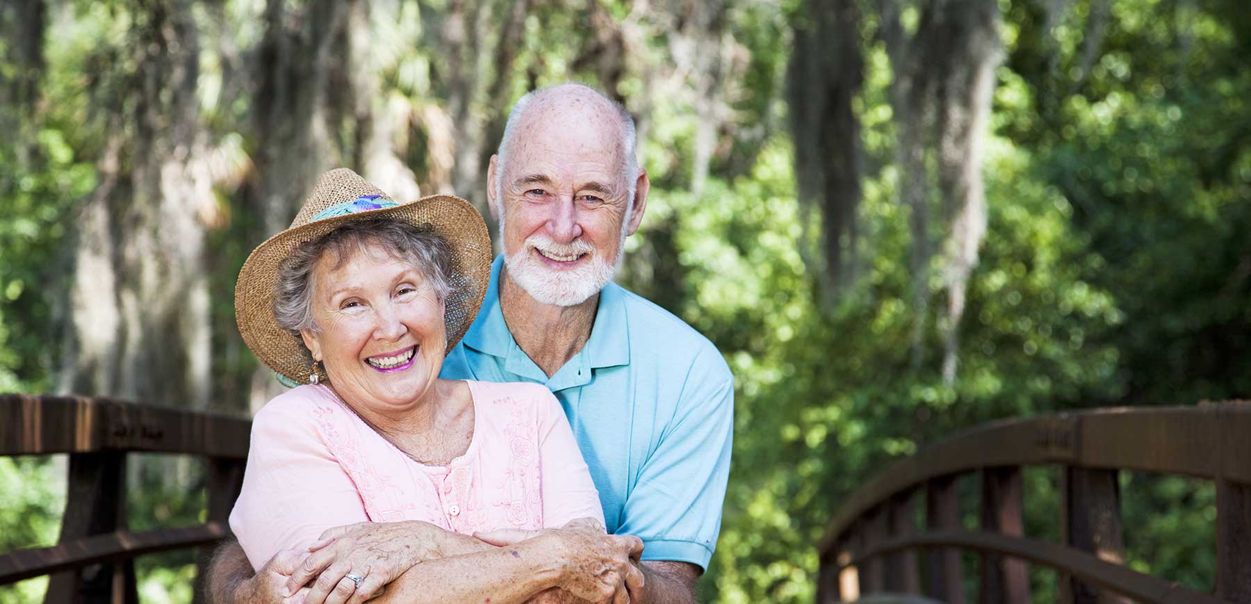 A photo of a senior couple on a bridge in Brunswick, GA. In the background we can see Spanish moss hanging from a live oak. The woman wears a sun hat and a pink sun dress. The man is also sharply dressed in a blue polo and khaki pants. Under the hot Georgia sun, the embrace, smiling directly at the viewer.